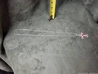 Silver chain necklace laid flat with a silver cross pendant that has red stones, chain length measured with a tape measure approx 12-15 inches.