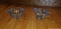 Front view of vintage blue glass creamer and sugar bowl on wooden floor showing scalloped edges and ribbed vertical pattern.