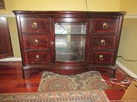 Front view of the dark reddish-brown wooden cabinet with a glass door and six drawers with brass ring handles on a patterned rug.
