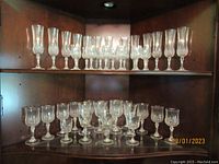 Wide view of upper and lower shelves showing 13 champagne flutes on top shelf and 27 matching cordial and wine glasses on lower shelf, plus some unmatched glasses