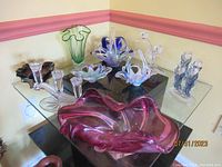 Wide view of vintage estate art glass items on glass table, showing various colored pieces including a large pink bowl in the foreground and other glass decorative items arranged around it.