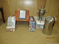 Wide view of all lot items including samovar, Kindle on wooden bed tray, Schlage knobs, small white container, metal trash bin, and paper towels.