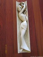 Frontal view of mid century chalkware wall sculpture showing the woman holding an ewer, with elongated stylized form and woodgrain backplate.