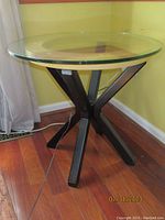 Side view of modern round side table with transparent glass top and black intersecting metal legs on wood floor.