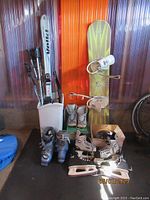 Several pairs of ski poles, skis, snowboard with bindings, ice skates, and boots arranged against a wall