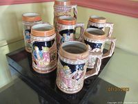 View of all seven ceramic beer steins arranged together showing their varied colorful historic and traditional designs with detailed reliefs and ornate handles.