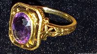 Close-up detail of the purple faceted amethyst or glass stone set in a 14K gold ring with decorative edges and engravings on the band.