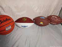 Four sports balls displayed on cloth background including two indoor I.F.L. footballs signed, plus an orange Baden basketball and a commemorative NFL football.
