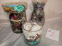 Three large glass jars filled with assorted costume jewelry shown from front and angle, highlighting variety of bead necklaces and metal pieces