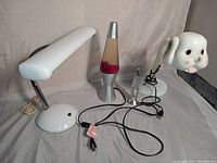 Three lamps: white metal desk lamp with gooseneck, silver and red lava lamp, ceramic puppy lamp head. All with cords arranged front center.