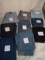 Top-down view of nine pairs of used jeans folded with handwritten size notes. Shows variety of blue, black and grey jeans with visible wear and labels.
