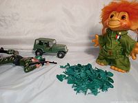Full lot view showing large DAM Things Army Troll doll in green outfit with orange hair, crawling shooting toy soldier in camouflage, olive green army jeep, and pile of small green army men figures.