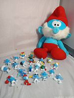 Large blue plush Papa Smurf with red hat and pants sitting, surrounded by 21 small Smurf plastic figurines in various poses