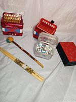 Full lot showing two red Bakelite accordions, wooden mallet, new drumsticks, Thomas Kincaid music chime box, and decorative Chinese box.