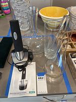 Photo showing the rechargeable black Cuisinart electric wine opener placed on charger base next to clear glass round-based wine carafe and a tall gooseneck decanter. Yellow and white bowl in background.