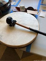 Photo showing hand drum with natural skin head and wooden mallet placed on top, small decorative candle and wooden dish holder visible in background.