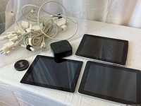 Three Apple iPads, Apple TV box, wireless charging pad, and collection of AC adapters and cables on a table.