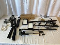 Overview photo showing two LED panel lights on stands, multiple tripods and selfie sticks, including foam grip and flexible leg tripods, arranged on a white surface.