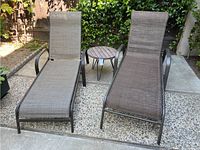 Two adjustable chaise lounges with metal frames and brown vinyl fabric outdoor seating next to wood and metal round side table on pebble concrete patio, showing overall lot composition and condition.