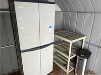 Wide shot showing all three items: white enclosed storage cabinet, beige plastic shelf unit, and stainless steel trashcan with black lid.