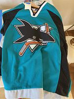 Front view of teal hockey jersey with large San Jose Sharks logo on the front.