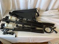 Four black light stands with two attached spiral CFL bulbs, black photography umbrellas, and soft box covering folded on white cloth surface.