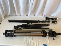 Three tripods laid out on a white cloth, showing their overall size and structure. One tripod is black and labeled GSX Ultra 45TM, folded compactly. Another tripod has extended silver legs and a phone mount attached. A webcam is visible on one tripod in another image.
