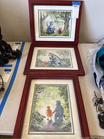 Three framed prints arranged vertically on a table, each featuring scenes with a child and a blue dinosaur in natural settings.