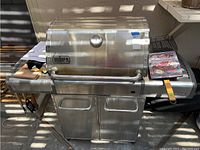 Front view of Weber Genesis stainless steel propane gas grill showing lid with thermometer, double-door storage below, side shelves with accessories and cookbook on top