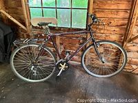 Side view of complete Raleigh bicycle showing frame, tires, saddle, pedals, and rear luggage rack in a wooden interior.
