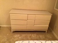 Full front view of white 7-drawer dresser against a wall with patterned wallpaper and carpeted floor.