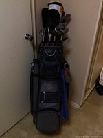 Full view of the black golf bag standing upright filled with golf clubs. Several pockets and compartments are visible. One wood club headcover marked "Jazz" and another similar headcover are present. Bag is visibly dirty and worn.