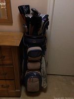 Full view of blue golf bag with multiple beige pouches and a variety of golf clubs inside.