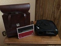 Photo of Kodak Carousel slide projector, brown leather carrying case, and instruction manual on wooden table.