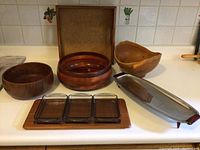 Photo shows various household items including wooden bowls, glass dishes on a wooden tray, and a metal soap dish with wooden soap all placed on a kitchen counter.