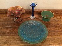 Four vintage glassware items on a beige background - pink ruffled bowl, blue hobnail plate, green pedestal dish, blue and white decorative vase