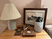 Wide view of all items on wooden table, showing ceramic lamp, jug, framed print behind, wooden box, stamps, and a textile item.