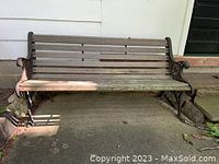 Front view of a used outdoor bench attached to concrete porch showing wrought iron armrests and wooden slats with evident wear and discoloration.