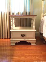 Full front view of the Vilas Canada side table beside a bed and window with curtains, showing drawer, shelf, turned column details, and wooden top.