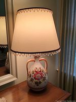 Front view showing the ceramic base with floral painting and fabric shade with decorative trim.
