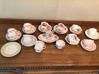 Photo of 11 assorted vintage bone china teacups with matching saucers arranged on wooden table. Various floral patterns and one plain pink set visible. Used condition with crazing and wear marks seen.