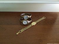 Photo showing two silver-tone stretch band watches and one gold-tone linked bracelet watch on a wooden surface.