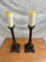 Pair of antique cast-iron candleholders each holding a white pillar candle on a wooden surface against a draped cloth background.