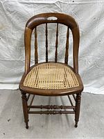 Front-facing photo of an antique wooden chair with a cane seat, showing the rounded top rail with cutout, turned spindles and legs.