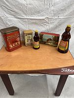 Photo showing five antique containers on a wooden table against a white fabric backdrop. From left to right: Imperial Blend India Tea tin, Wilbur's Cocoa tin, small Aunt Jemima syrup bottle, decorative rural landscape tin box, large Aunt Jemima syrup bottle.