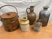 Photo of antique wooden ice bucket and four assorted pottery items on wooden table against white fabric background.
