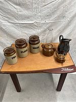 Photo of six ceramic kitchen items on a wooden surface: four storage jars with lids labeled Tea, Flour, Sugar, one unlabeled, two brown teapots, and one black pitcher.