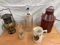 Four vintage containers arranged on a white cloth: a metal lantern, a crystal oil lamp, a ribbed glass seltzer bottle with siphon top, and a ceramic pitcher with flowers