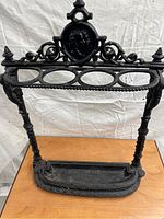 Front view of black cast iron umbrella stand with ornate details including scrollwork, figure roundel, and umbrella holder slots