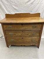 Front view of antique wooden dresser with three drawers showing wood grain, brass handles, scalloped base and carved back panel.